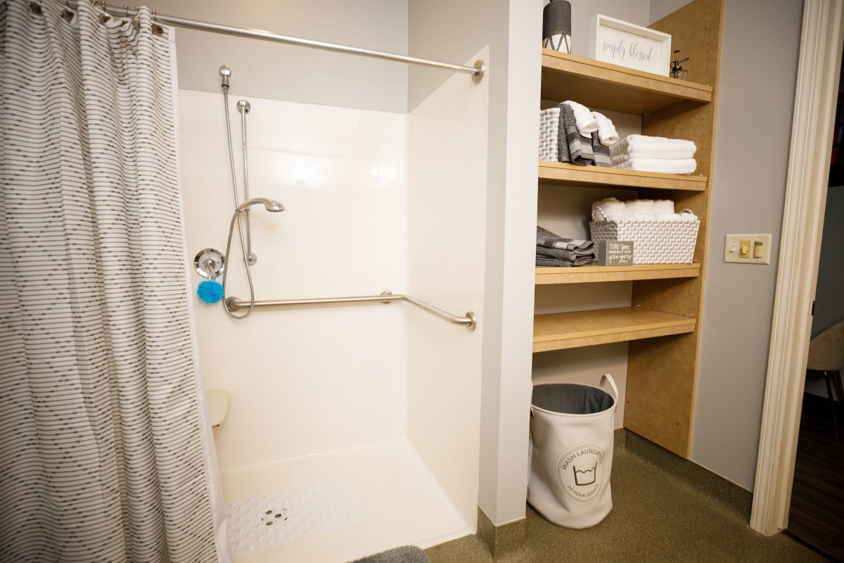 A bathroom with a walk-in shower equipped with grab bars, adjacent shelving holding towels and toiletries, and a small trash bin.