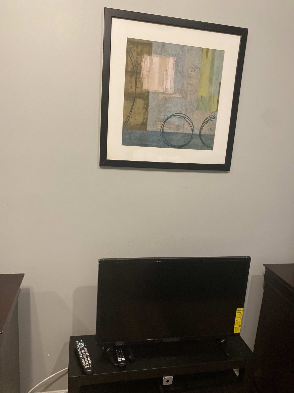 A framed abstract painting hangs on the wall above a television set placed between two pieces of furniture.