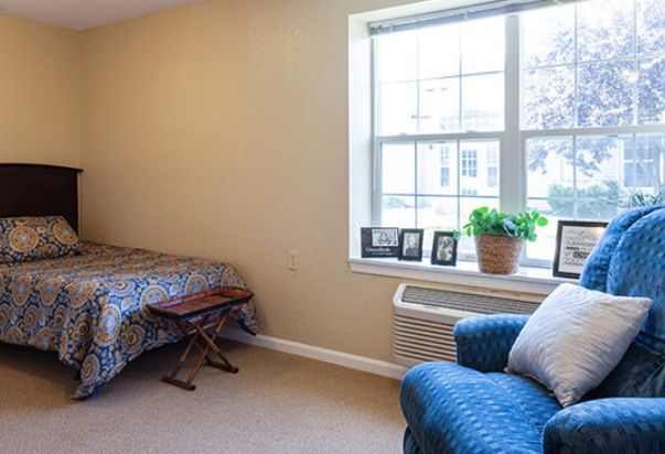 A cozy bedroom with a bed, a blue chair, and a window with potted plant decor.