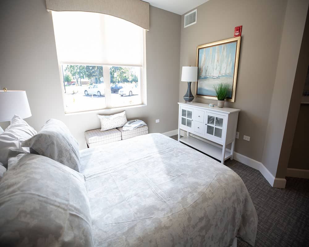 A cozy bedroom with a large window, featuring a neatly made bed, dresser with a lamp, and wall art.