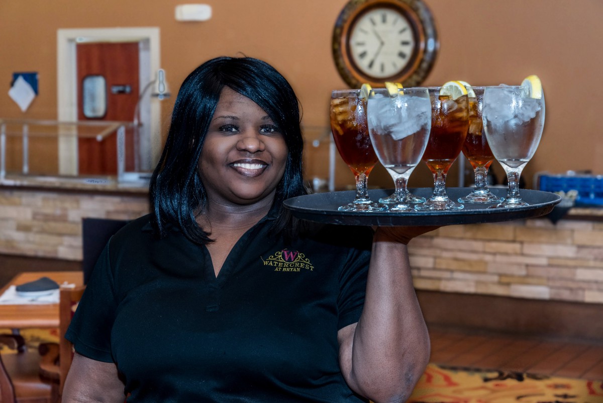 A smiling waitress holding a tray of iced tea glasses in a restaurant setting.