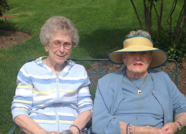 Two elderly women sitting on a bench outdoors, one wearing a sun hat and the other wearing glasses.