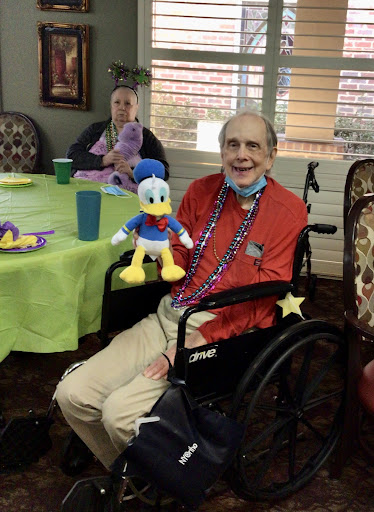 An elderly man in a wheelchair happily holds a plush Donald Duck toy during a social gathering at a residential facility.