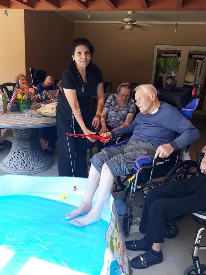 An elderly person relaxes with their feet in a small pool, assisted by a caregiver, as others enjoy the moment at a care facility.