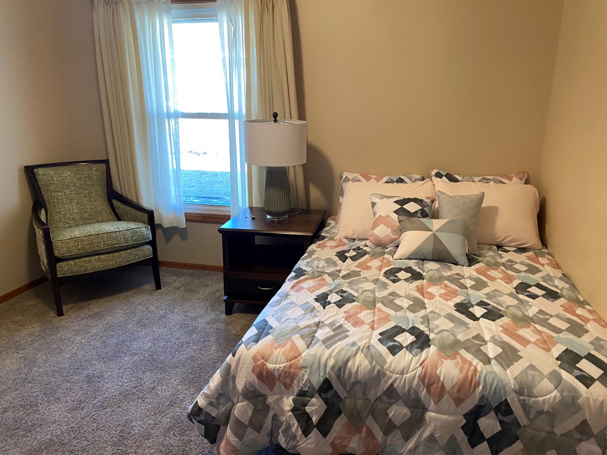 A cozy bedroom featuring a neatly made bed with patterned bedding, a nightstand with a lamp, and a window with curtains.