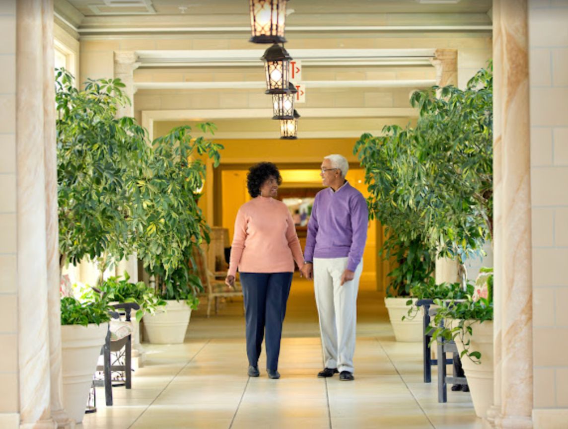 An elderly couple walking hand in hand through a plant-lined corridor.