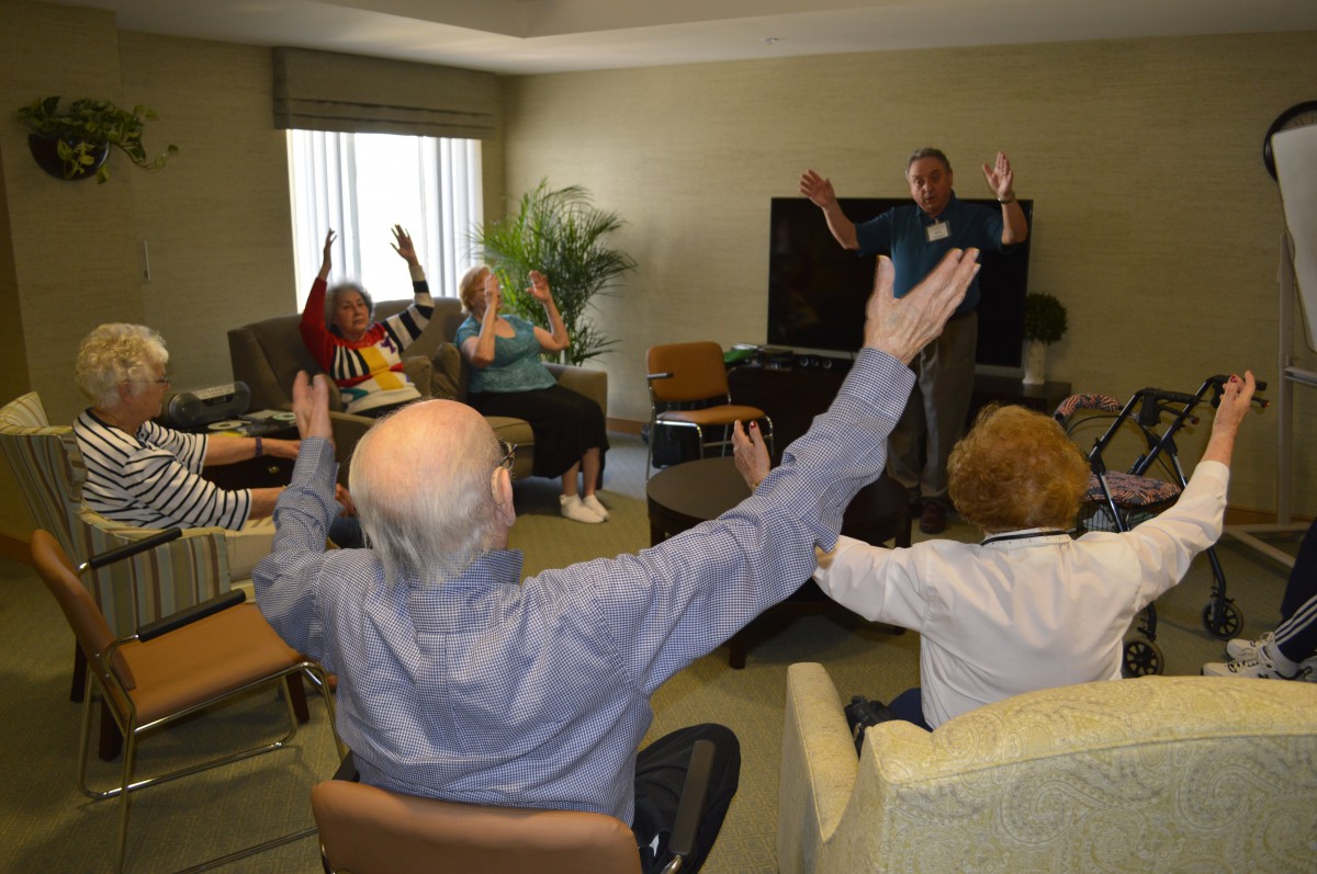 A group of elderly individuals participating in an indoor exercise session, raising their arms as part of the activity.
