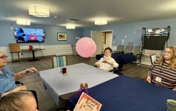 An elderly woman sitting in a recreation room, playing with a pink balloon over a table.