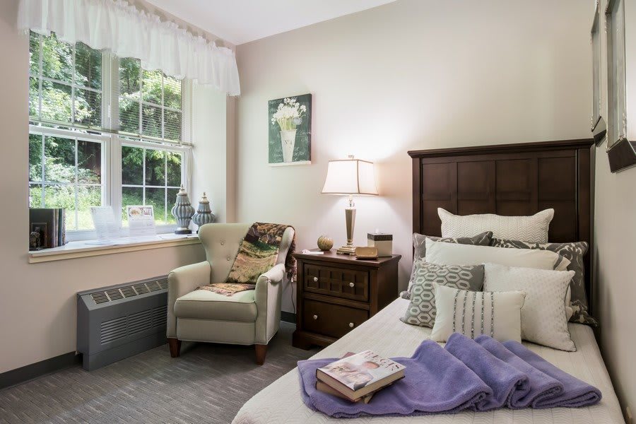 A cozy bedroom with a neatly made bed, comfortable armchair, and a side table with a lamp.