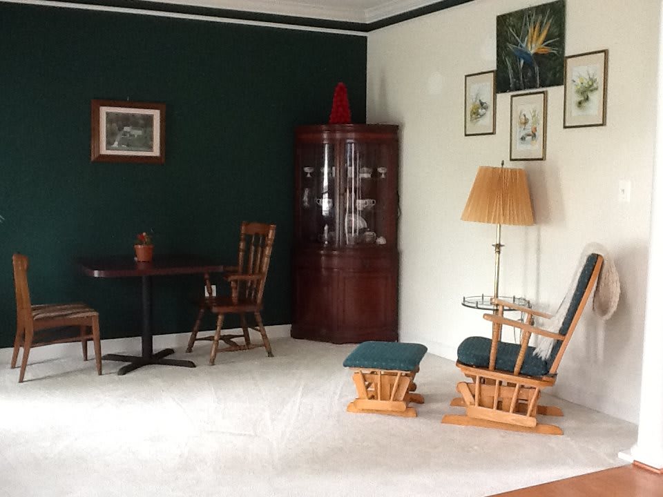 A cozy living room with dark green and white walls, featuring wooden furniture including chairs and a table, a cabinet, floor lamp, and various wall decorations.