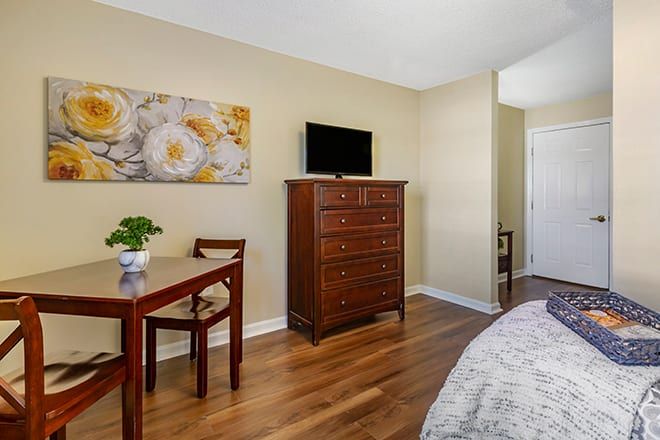 A cozy bedroom featuring a wooden dresser with a TV on top, a small table with chairs, floral artwork, and a potted plant on a wooden floor.