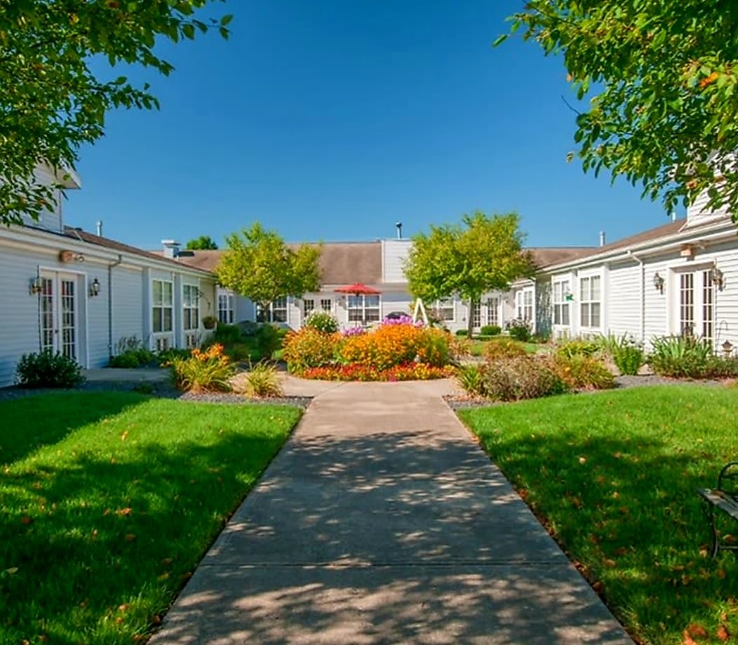 A scenic view of a garden pathway leading to a building, surrounded by lush greenery and vibrant flowers under a clear blue sky.