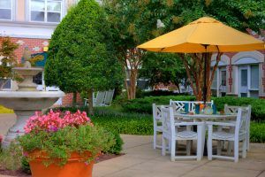 A serene patio setting with a table, chairs, and a yellow umbrella surrounded by lush greenery and colorful flowers.