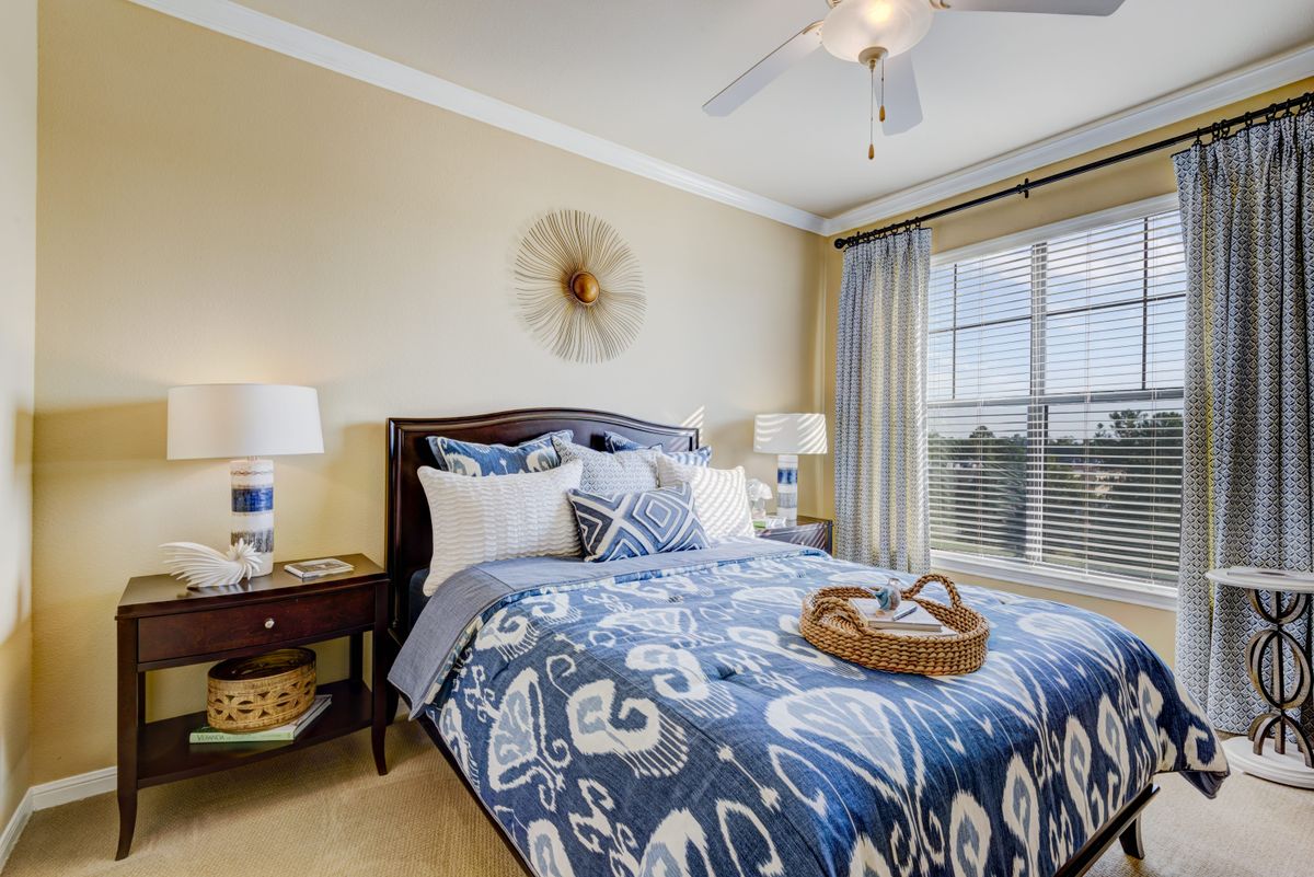 A cozy bedroom featuring a bed with blue patterned bedding, flanked by two nightstands with lamps, and a ceiling fan above. Natural light streams in through a large window, enhancing the room's inviting atmosphere.