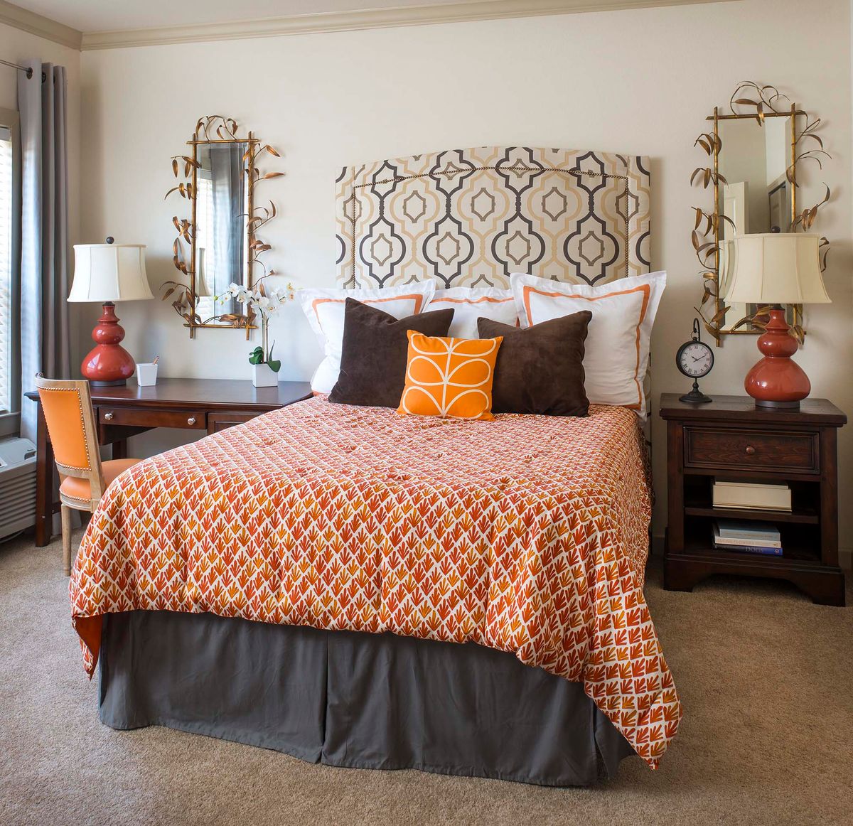 A well-decorated bedroom featuring a bed with orange patterned bedding, flanked by two bedside tables with matching lamps and ornate mirrors.
