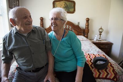 An elderly couple smiling and sitting together in a cozy bedroom.