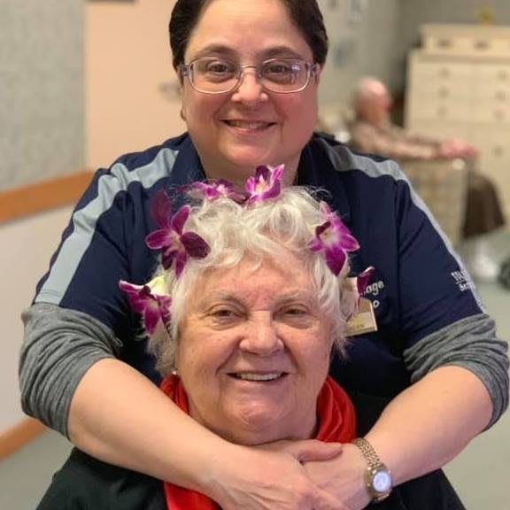 A smiling elderly woman with flowers in her hair, embraced by a younger person indoors.