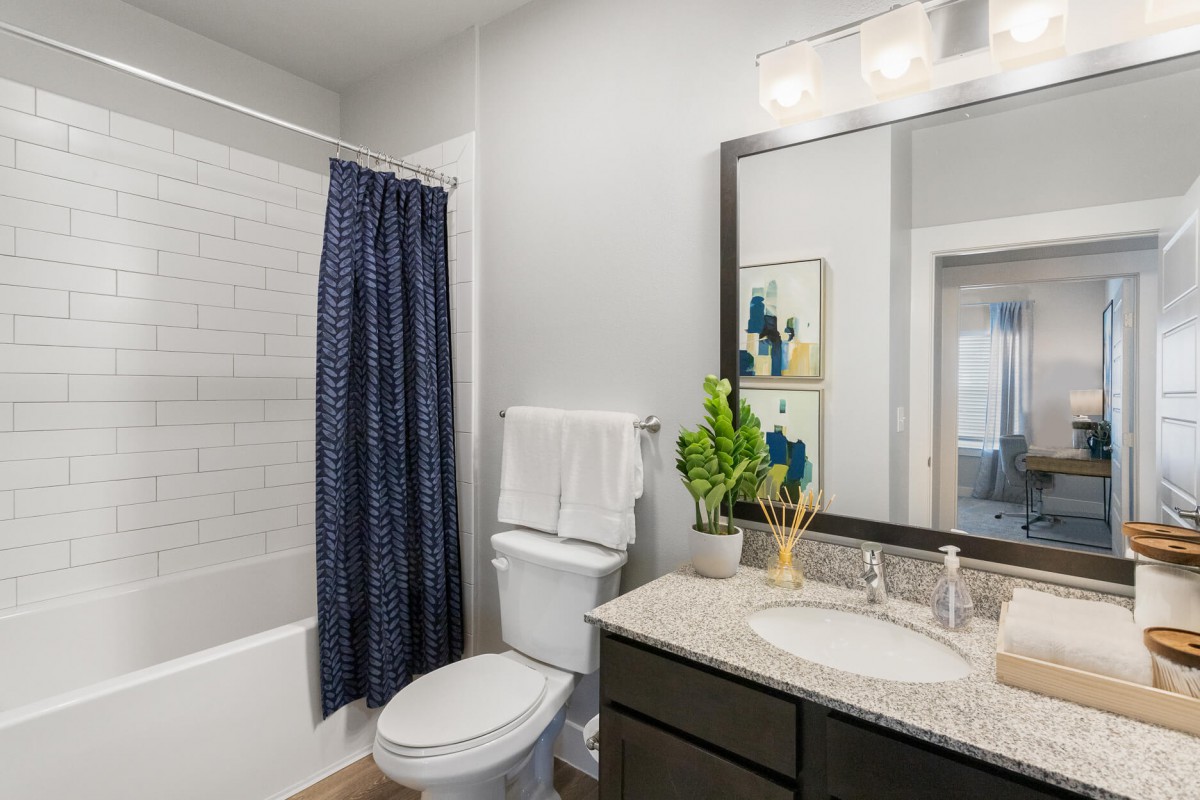 A modern bathroom featuring a shower with a blue curtain, a toilet, and a sink with a mirror above. The decor includes a potted plant and indoor lighting.