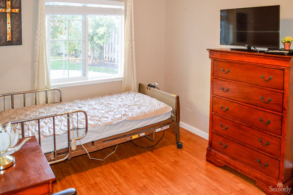 A bright bedroom with a single hospital bed next to a window, featuring a wooden dresser and a TV.