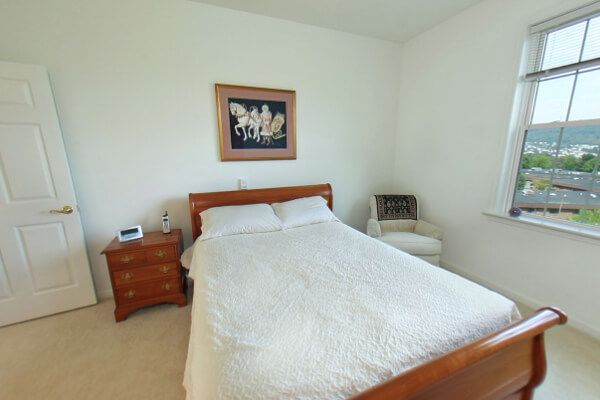 A minimalist bedroom with a neatly made bed, wooden nightstand, artwork on the wall, and a chair in the corner.