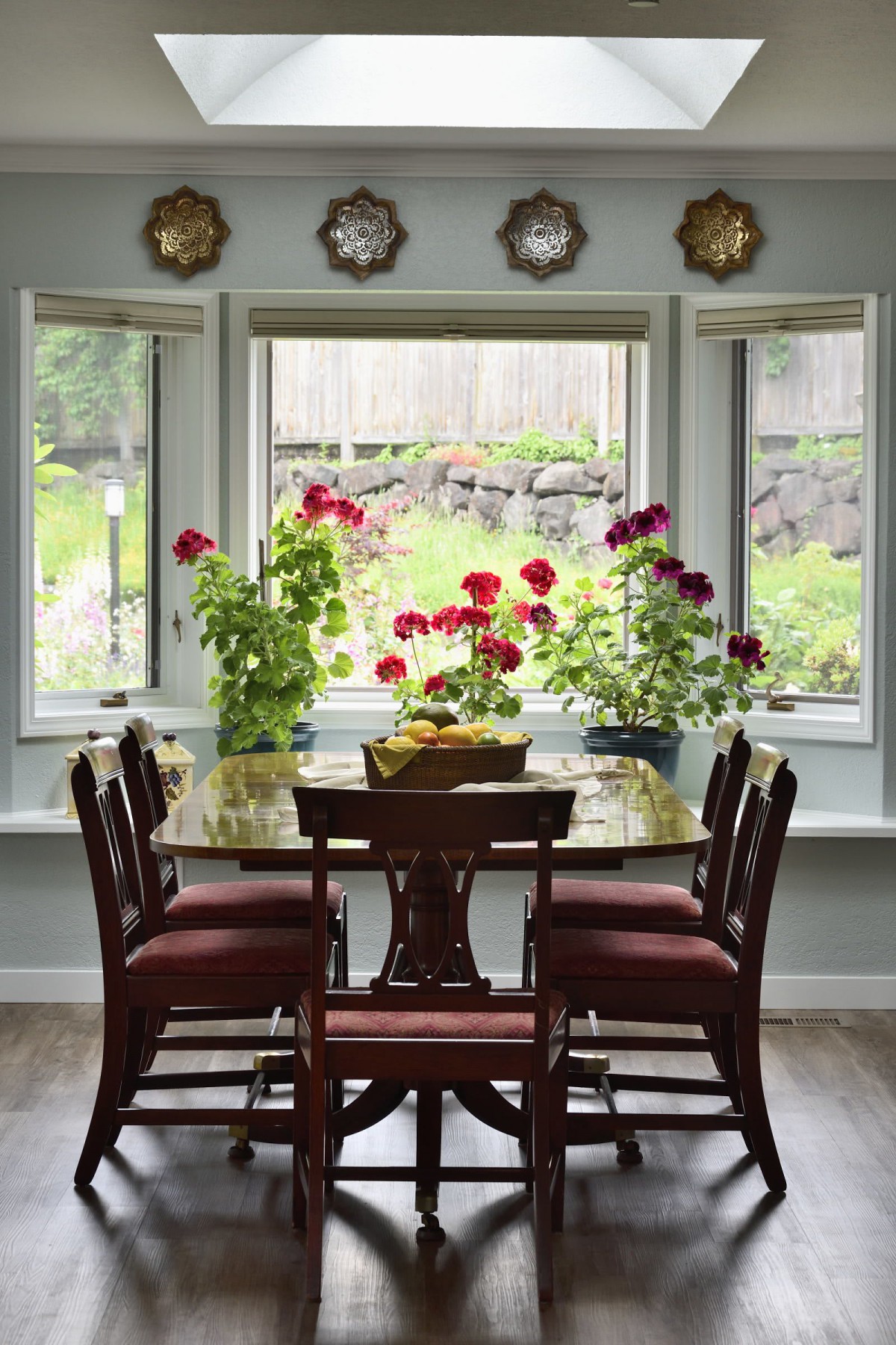 A cozy dining room with a wooden table and chairs, surrounded by large windows and vibrant potted flowers.