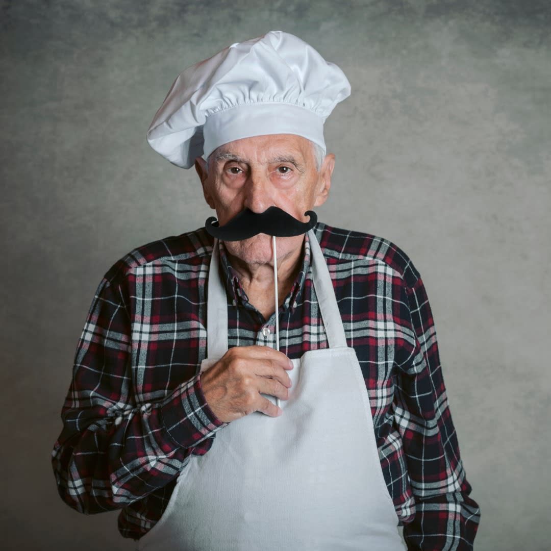 An elderly man dressed as a chef with a large mustache.