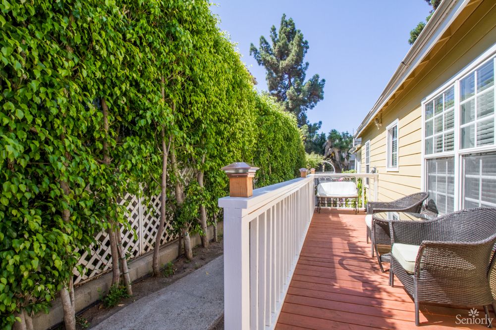 A sunny deck alongside a house with lush greenery and a white fence.