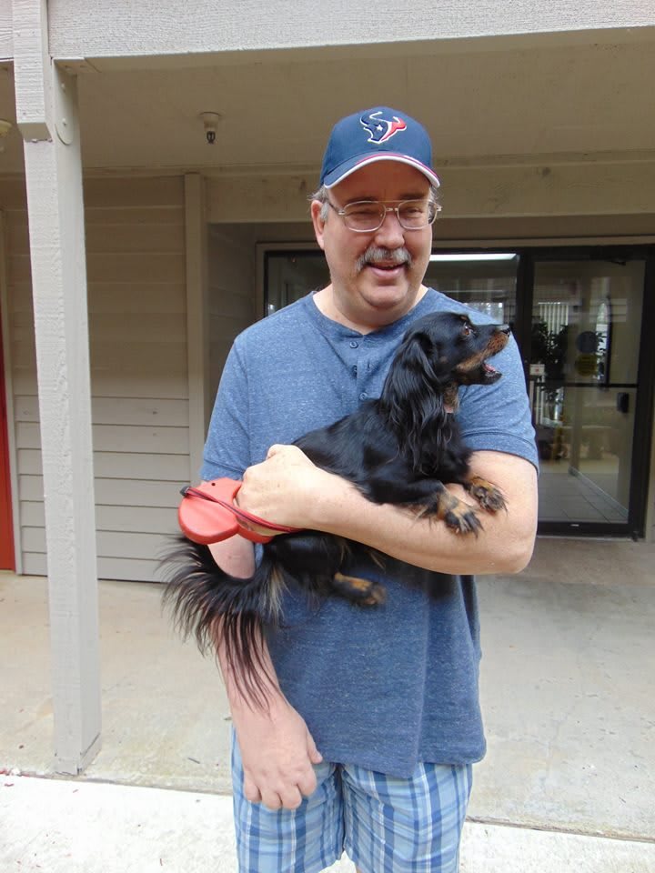 A man wearing a hat and blue shirt, holding a small black dog indoors.