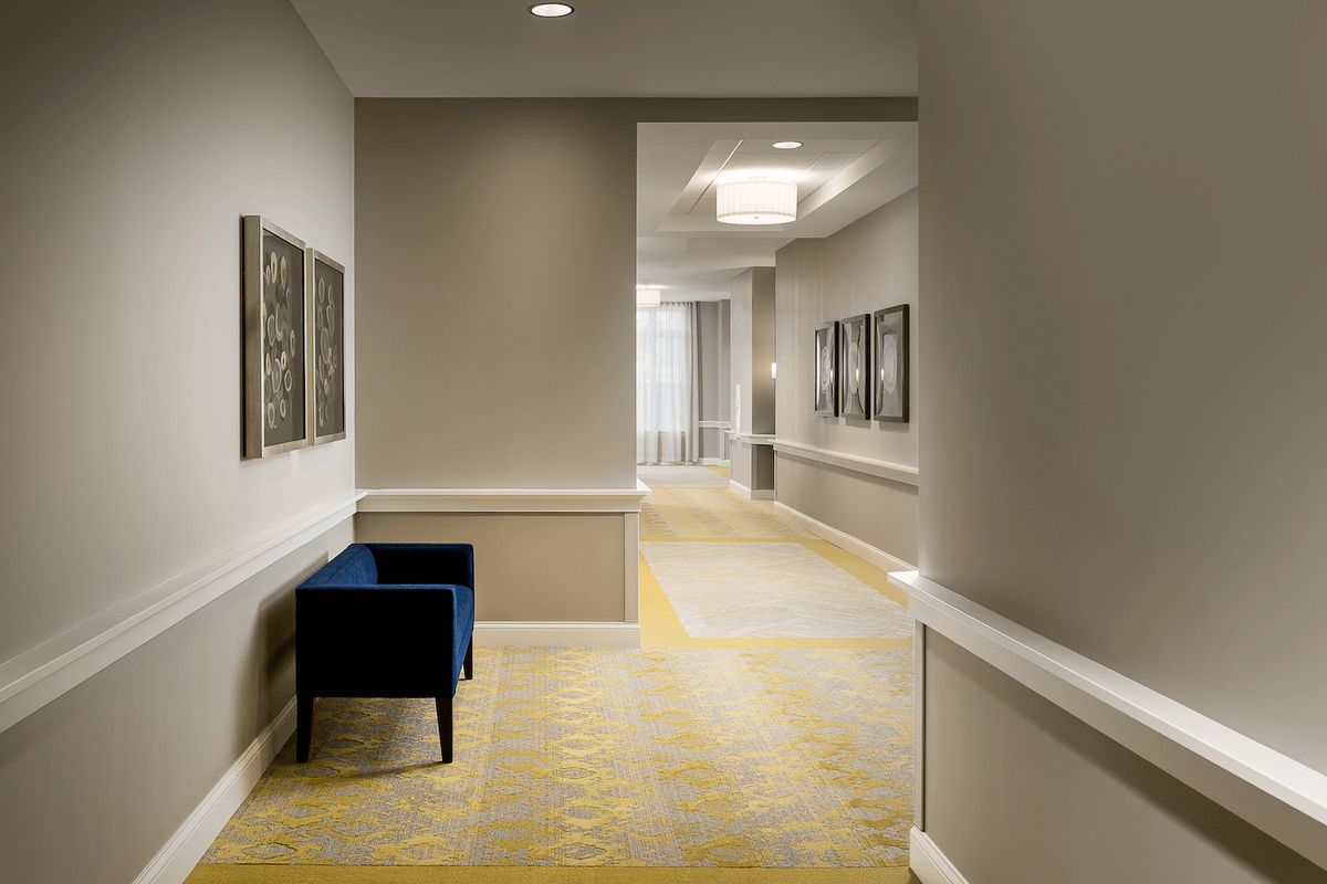 A modern hotel hallway with artwork on the walls and a blue chair against a beige backdrop.