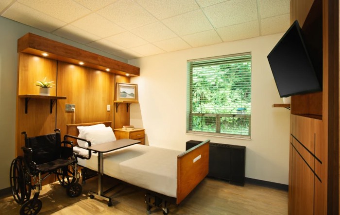 A hospital room with a patient bed, wheelchair, and a window with a view of the outdoors.