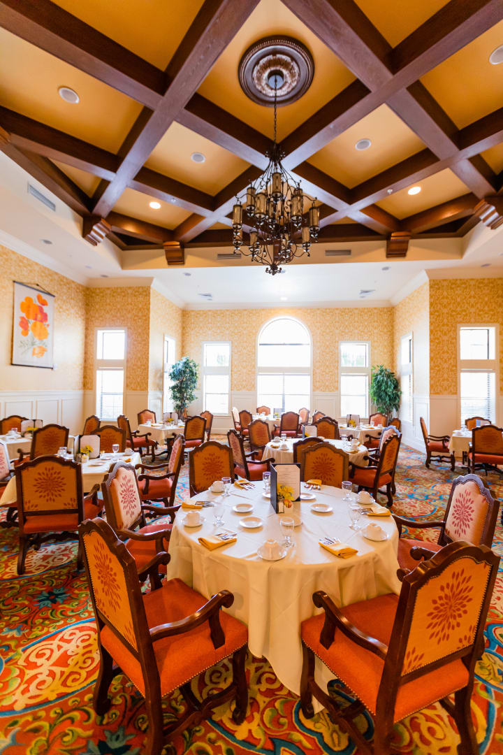 An elegant dining room with round tables, plush chairs, and a chandelier under a coffered ceiling.