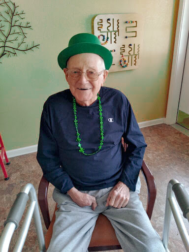An elderly man wearing a green hat and necklace, celebrating in a cheerful indoor setting.