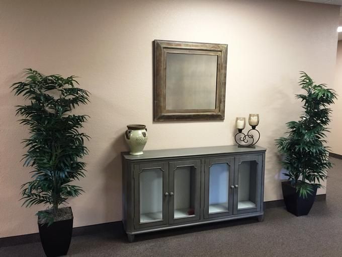 A neatly decorated office corner with a wooden cabinet, framed mirror, and potted plants.