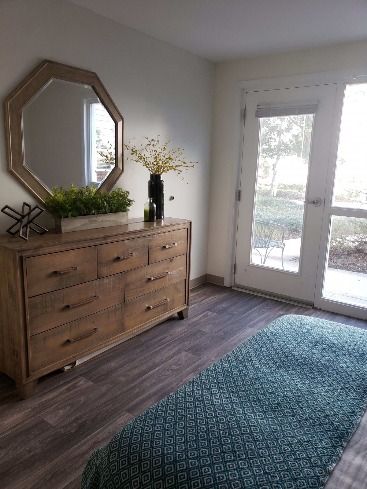 A well-lit bedroom featuring a wooden dresser with a decorative mirror and plants, alongside a blue rug and large windows.