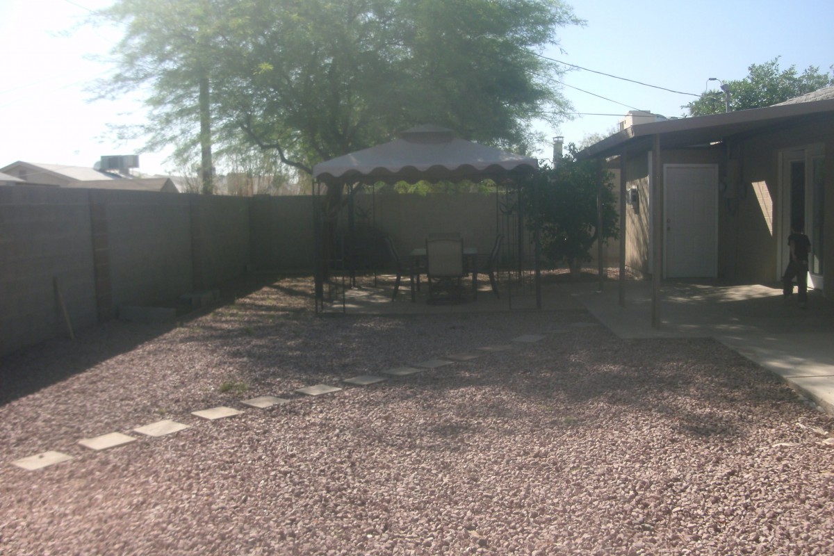 A sunny backyard with gravel ground, featuring a gazebo and outdoor seating area surrounded by trees.