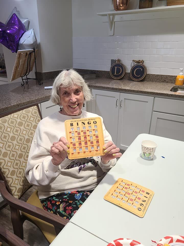 An elderly woman smiling while holding a bingo card at a table.
