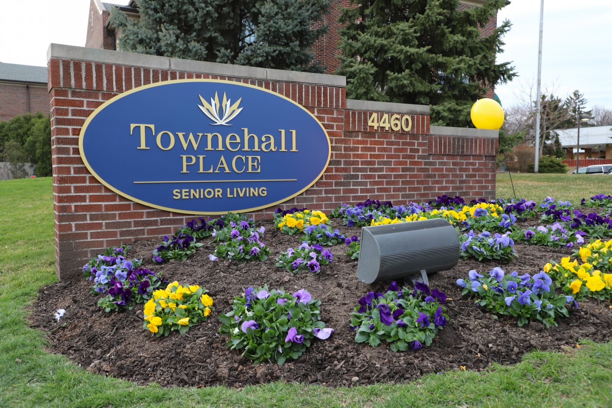 A sign for Townehall Place Senior Living surrounded by colorful flowers and a small balloon tied to the signpost.