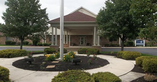 A building entrance with a covered porch, surrounded by neatly arranged landscaping and a flagpole.
