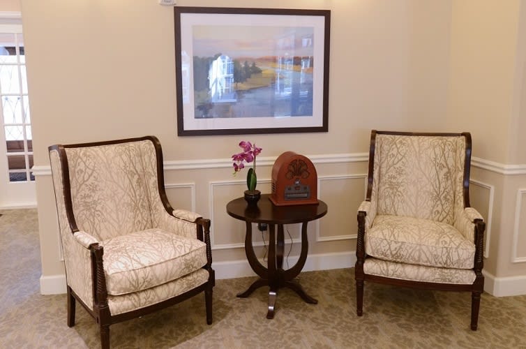 A cozy sitting area featuring two upholstered chairs, a small wooden table with a vintage radio and flowers, and a scenic painting on the wall.