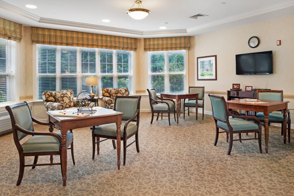 A cozy and well-lit living and dining area with multiple tables and chairs, featuring large windows and warm decor.