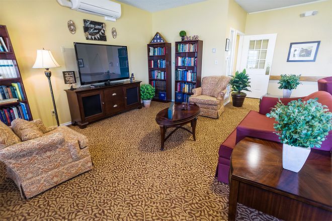 A cozy living room featuring a large TV, armchairs, bookshelves, and a coffee table on a patterned carpet.