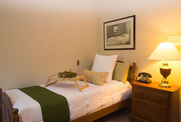 A cozy bedroom with a single bed featuring green accents, a nightstand with a lamp, and a decorative tray.