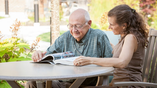 An elderly man and a woman enjoy reading together outdoors, sharing smiles over a magazine.