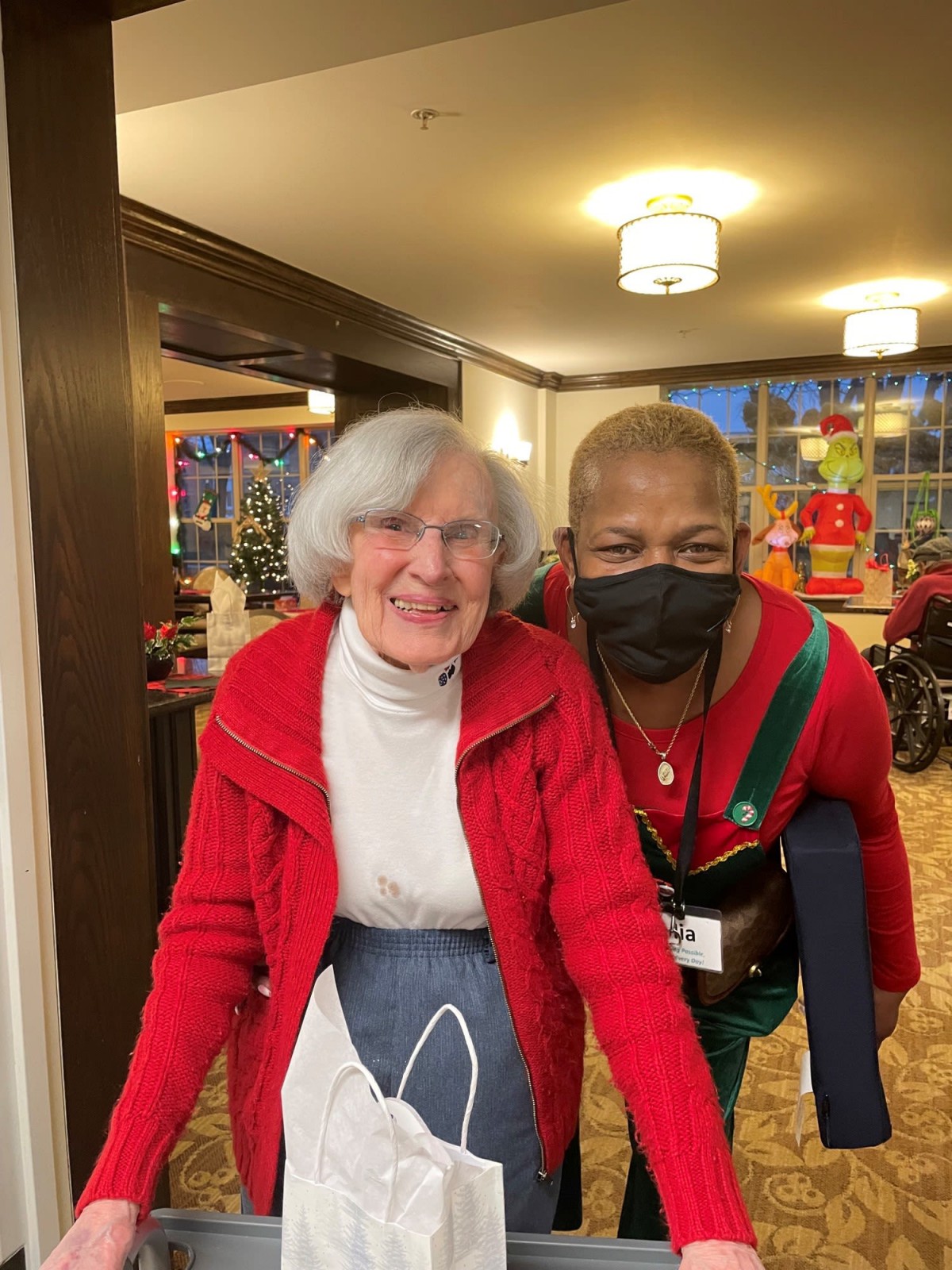 An elderly woman and her caregiver share a moment together indoors, both wearing red clothing.