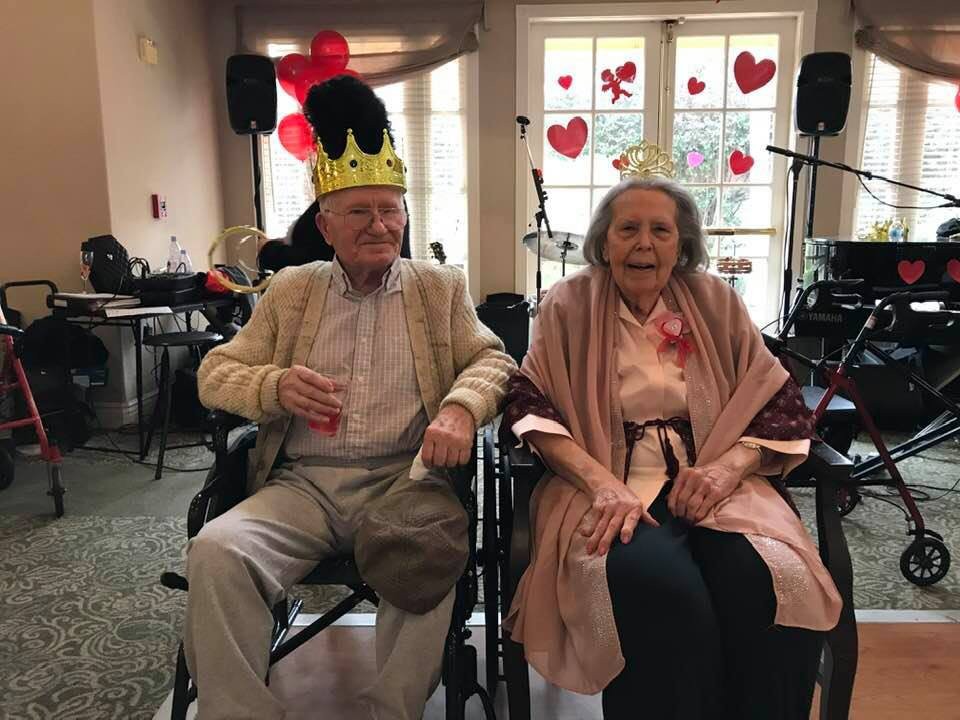 An elderly couple wearing crowns, sitting together at a celebration with heart decorations in the background.