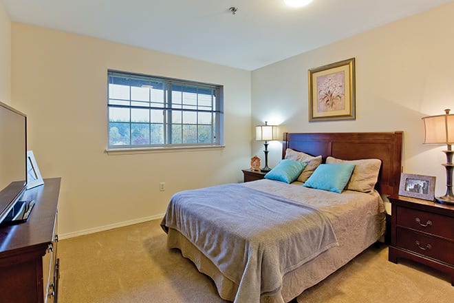 A cozy bedroom with a neatly made bed, side table with a lamp, and a window providing natural light.