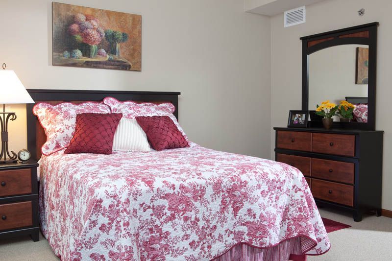 A cozy bedroom featuring a bed with floral-patterned bedding and pillows, alongside a wooden dresser with a mirror.