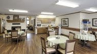A cozy dining area with several tables and chairs, connected to a kitchen with wooden flooring and warm lighting.