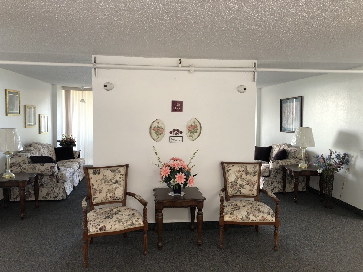 A living room seating area with two decorative chairs and a small table adorned with flowers, set against a white wall with framed artwork.