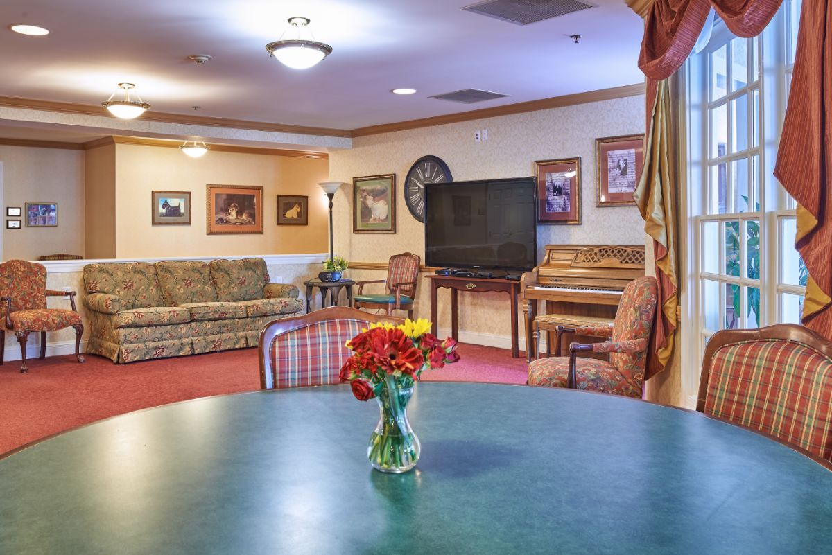 A cozy living room with a table, couch, television, and piano, decorated with flowers and artwork.